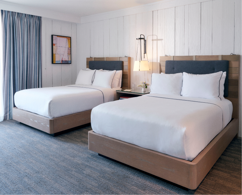 Two well-arranged cozy beds, separated by a side table with a glowing lamp above it, and a portrait mounted on the left wall.