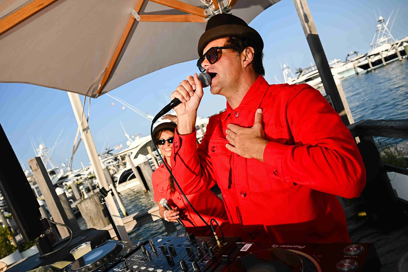 DJ performing with microphone at waterfront deck with marina in background