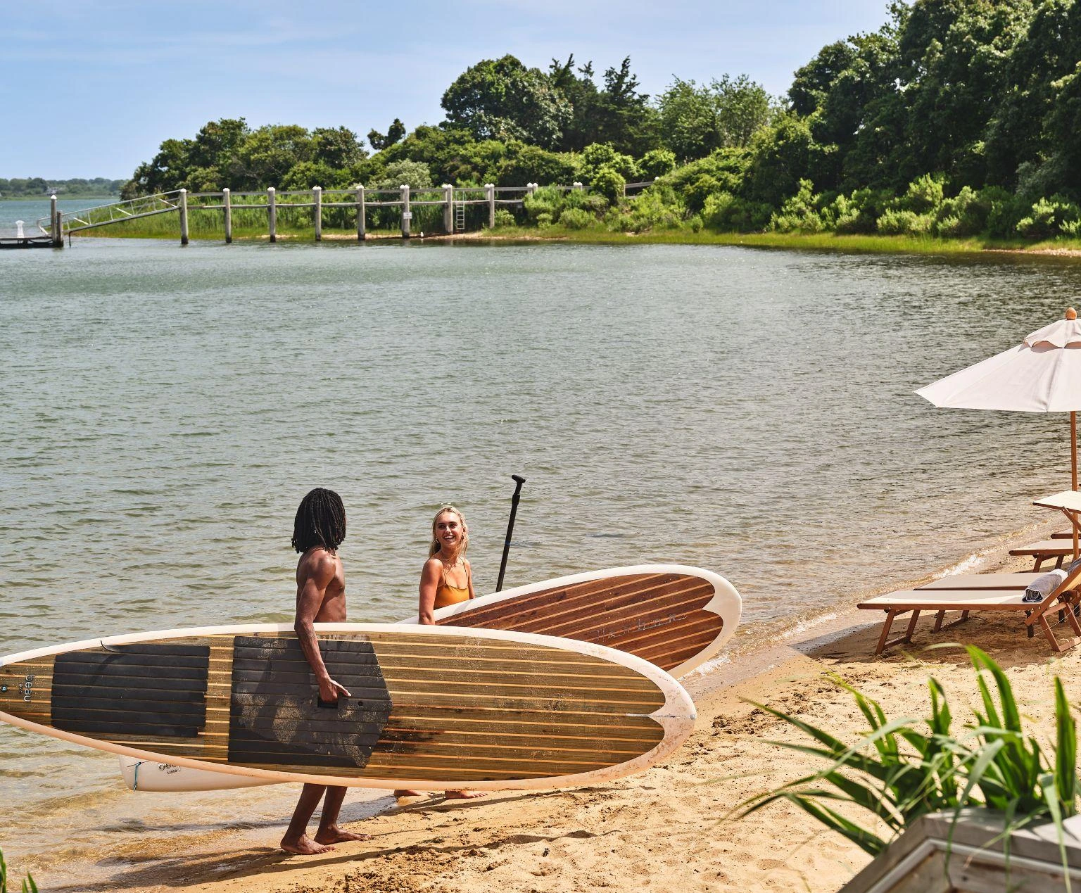 Two people carrying paddleboards along a sandy lakeshore near lounge chairs